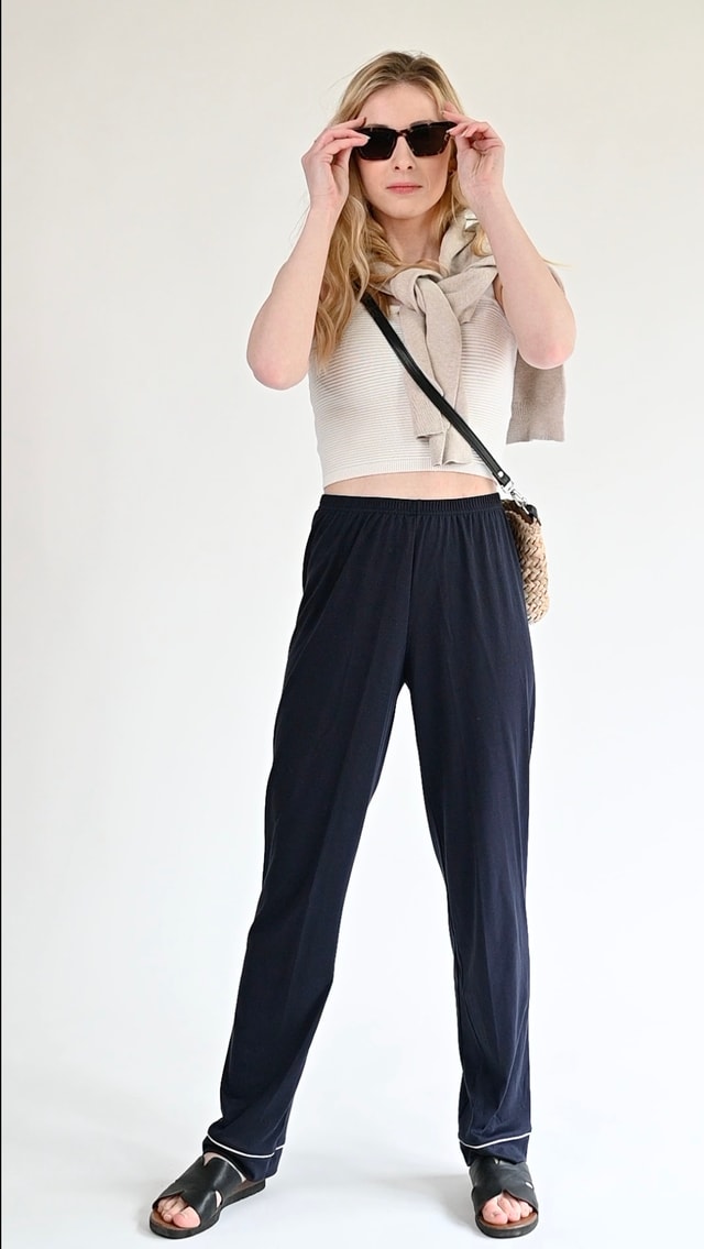 Eine Frau trägt eine luftig-leichte Pyjamahose von CALIDA, ein Tanktop, eine Basttasche im Boho-Stil, offene Schuhe und einen Pullover um die Schultern.