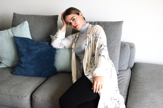 Frau sitzt auf grauem Sofa und trägt Kimono und Rollkragenpullover von CALIDA. Der Rollkragenpulli ist hellblau und der Kimono cremefarbig.
