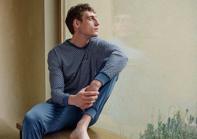 Mann trägt blauen Pyjama von CALIDA. Er sitzt in einem Haus neben einem grossen Fenster.