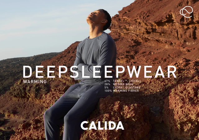 Man in dark blue sleepwear sits on reddish rocks in a volcanic landscape and enjoys the sun with his eyes closed. Text overlay with ‘DEEPSLEEPWEAR’, material composition and the CALIDA brand.
