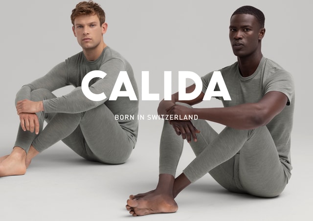 Zwei Männer sitzen auf dem Boden in graugrüner Loungewear, einer trägt ein langärmeliges Oberteil, der andere ein T-Shirt. Beide schauen ernst in die Kamera. Großes Logo CALIDA mit dem Slogan Born in Switzerland im Vordergrund.