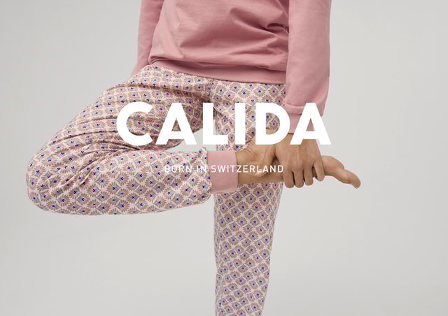 Person in pinkem Sweatshirt und gemusterten Pyjamahosen hält beim Yoga-Üben ihren Fuß, mit dem Markennamen CALIDA und dem Slogan 'Born in Switzerland' im Vordergrund.