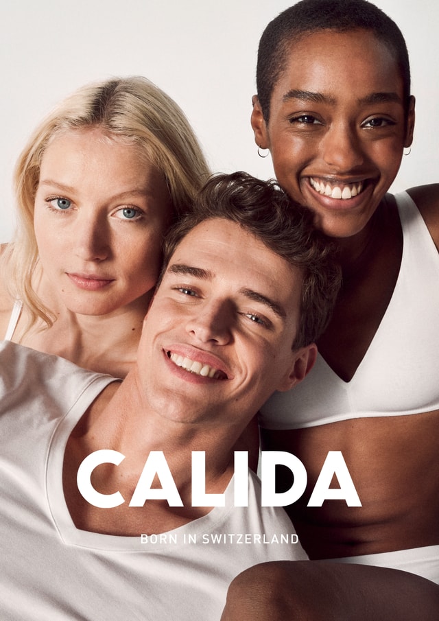 Drei lächelnde Models – eine blonde Frau, ein brünetter Mann und eine dunkelhäutige Frau mit Kurzhaarschnitt – tragen weiße CALIDA-Basics und posieren eng beieinander. Der Schriftzug 'CALIDA – Born in Switzerland' ist über dem Bild platziert.