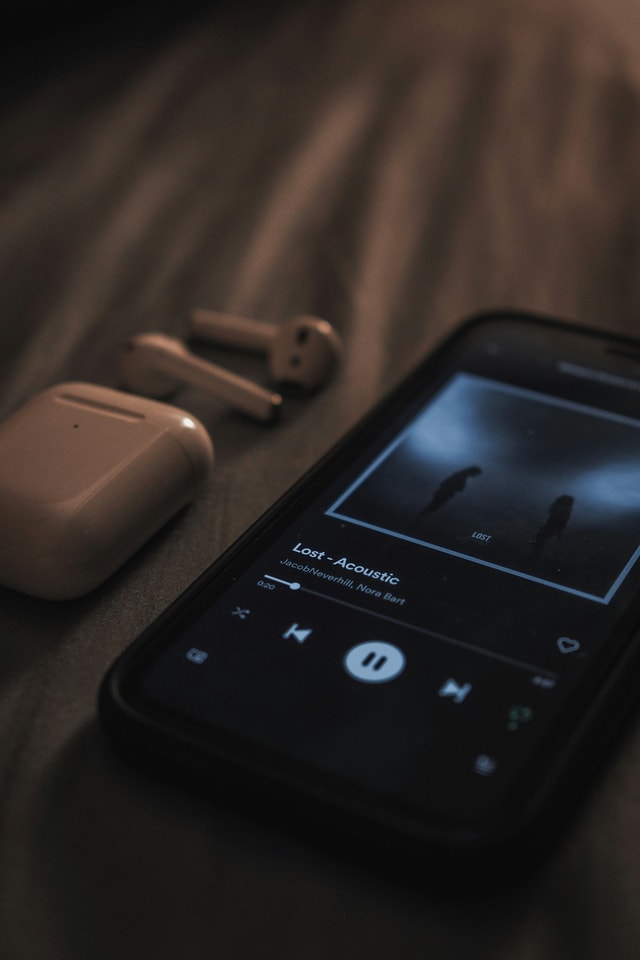 An iPhone with a music app open showing the song 'Lost - Acoustic' by JacobNeverhill and Nora Bart. Next to it lie wireless in-ear headphones (AirPods) and their charging case on a bed or soft surface.