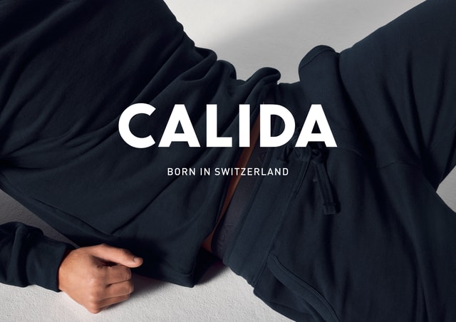 Person lies relaxed in dark loungewear, slightly raised top reveals the waistband of the trousers. Large ‘CALIDA’ logo with ‘Born in Switzerland’ underneath.