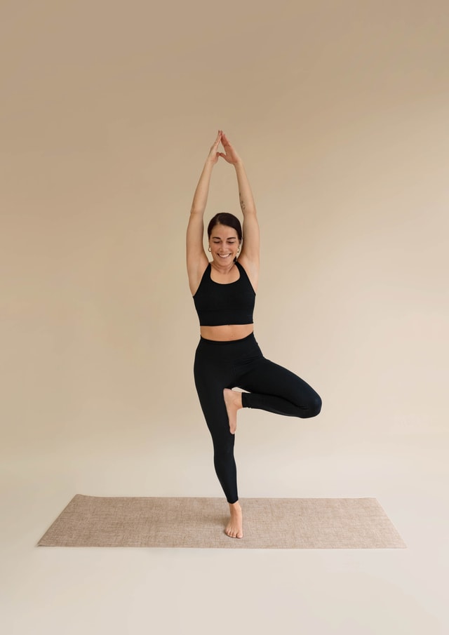 Woman wears black bustier and leggings from CALIDA. She does the yoga pose 'The Tree'.