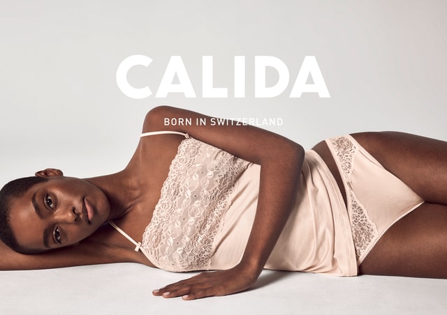 Dunkelhäutige Frau liegt seitlich auf einem hellen Untergrund, trägt ein zartrosafarbenes Spitzentop mit schmalen Trägern und eine passende Unterhose mit Spitzenverzierung. Großes Logo CALIDA mit dem Slogan Born in Switzerland im Vordergrund.