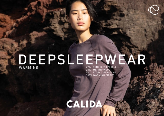 Advertisement for CALIDA DEEPSLEEPWEAR. A woman with Asian features wears a long-sleeved, dark purple sleep shirt made of TENCEL™, merino wool and LYCRA®. She poses in front of a rocky, volcanic landscape. The text describes the materials and the warming properties of the fabric.