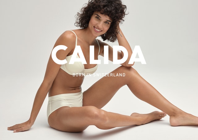 Smiling woman with curly dark hair sits on the floor in light-colored underwear, resting her head on one hand and looking directly at the camera. Large CALIDA logo with the slogan Born in Switzerland in the foreground.