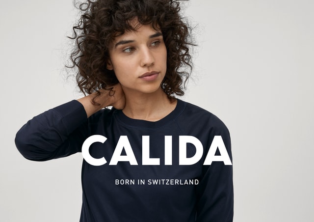 A woman wears a long-sleeved shirt by CALIDA from the Favourites Dreams range. She is holding her right arm at the back of her neck. The text 'CALIDA - Born in Switzerland' can be seen in the picture.